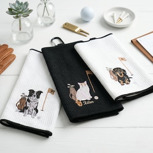 K&ouml;nnte beinhalten: Drei Golfhandt&uuml;cher mit schwarzen und wei&szlig;en R&auml;ndern. Jedes Handtuch hat ein gesticktes Design einer Golftasche, einer Fahne und eines anderen Tieres: einem Border Collie, einer Katze und einem Dackel. Die Handt&uuml;cher sind aus strukturiertem Material.