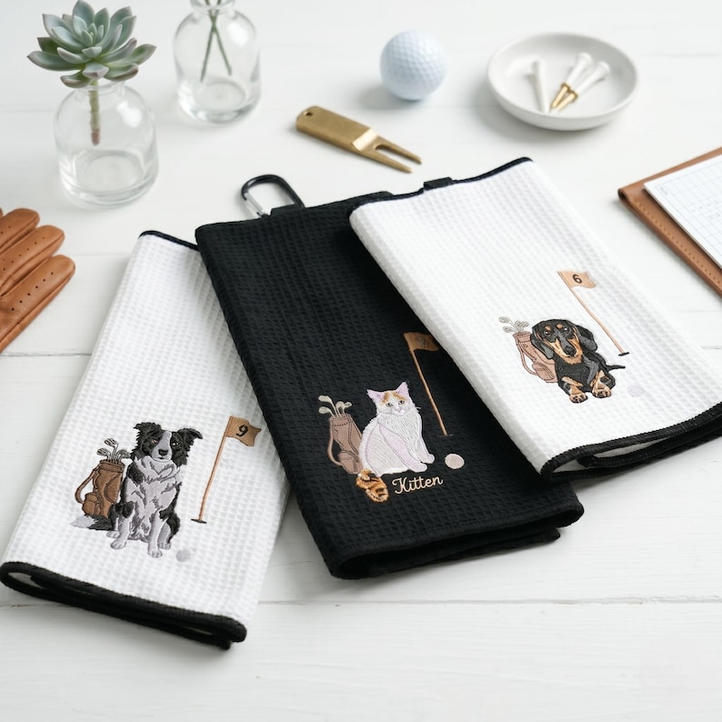 K&ouml;nnte beinhalten: Drei Golfhandt&uuml;cher mit schwarzen und wei&szlig;en R&auml;ndern. Jedes Handtuch hat ein gesticktes Design einer Golftasche, einer Fahne und eines anderen Tieres: einem Border Collie, einer Katze und einem Dackel. Die Handt&uuml;cher sind aus strukturiertem Material.