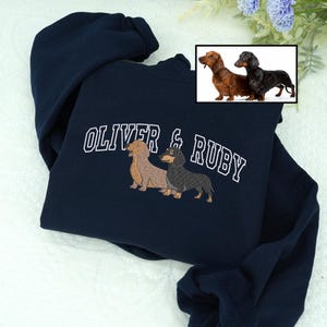 Navy embroidered sweatshirt featuring two dachshunds, one tan and one black-and-tan, with the names “OLIVER & RUBY” in varsity-style lettering. Personalized pet portrait embroidery showing realistic stitch details — a custom gift for dog owners and dachshund lovers.
