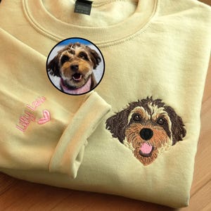 May include: A pale yellow crewneck sweatshirt featuring a detailed embroidered portrait of a brown and cream-colored dog on the chest. The sleeve is embroidered with the name Lily Lou in pink script lettering next to a small pink heart icon.