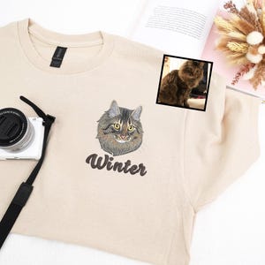 Custom embroidered sand sweatshirt featuring a lifelike portrait of a fluffy brown tabby cat with golden eyes, personalized with the name Winter stitched beneath in elegant script. Unique pet embroidery gift for cat moms, memorial keepsakes, and animal lovers. Perfect personalized clothing for birthdays, anniversaries, or Christmas.