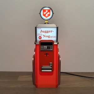 以下が含まれることがあります： 赤い銀色のビンテージスタイルのディスペンサーで、白い看板には「Jugger-Nog」と「本物の卵を使用」と書かれています。ディスペンサーの前面には赤い十字架があり、銀色のトップには赤い十字架があります。