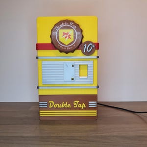 Puede incluir: Un dispensador de cerveza de doble grifo de estilo vintage amarillo y marrón con una franja roja y un letrero de 10¢. El dispensador tiene una ventana con una rejilla plateada y el texto "Double Tap" en la parte delantera.