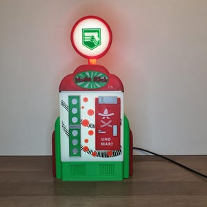 Peut inclure: Une pompe à essence rouge, blanche et verte en forme de distributeur automatique avec une lumière sur le dessus. La machine a une enseigne qui dit "Music Kick" et "Uno Mas!" avec un graphique de sombrero rouge et blanc.