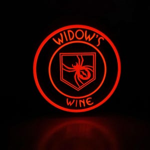 Peut inclure: Un emblème circulaire rougeoyant avec les mots "WIDOW'S WINE" et un graphisme d'araignée. L'araignée est à l'intérieur d'un bouclier. Le design est illuminé sur un fond noir.