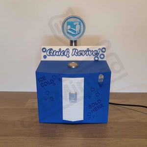 Peut inclure: Modèle 3D imprimé en bleu et blanc d'une machine "Quick Revive!" de la série de jeux vidéo Call of Duty. La machine a une pancarte qui indique "Ice Cold" et "Sold Here".