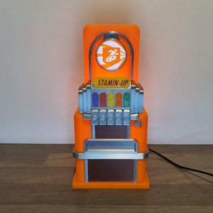Peut inclure: Un distributeur automatique de jouets orange et argent avec l'inscription "STAMIN-UP" sur le devant. La machine a une fenêtre en plastique transparent avec un logo blanc d'une personne courant. La machine a six boutons avec des lumières de différentes couleurs.
