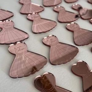May include: A collection of rose gold mirror acrylic dress-shaped place cards with names written on them. The place cards are perfect for a wedding or bridal shower.