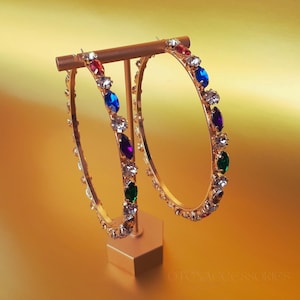 Puede incluir: Aretes de aro dorados con una fila de piedras preciosas de colores. Las piedras preciosas son rojas, azules, moradas, verdes y transparentes.