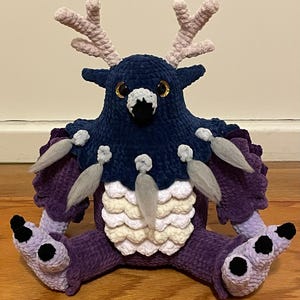 以下が含まれることがあります： 紫色の毛皮、白と灰色の鱗、そして角のある青い頭を持つ生き物の形をした、かぎ針編みで作ったぬいぐるみ。生き物は、大きな金色の目と黒い嘴をしています。