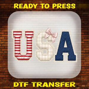 May include: A DTF transfer design featuring the letters U, S, and A in a varsity font. The U has red and white horizontal stripes, the S is beige with a light plaid pattern and a pink bow, and the A is navy blue with white polka dots.