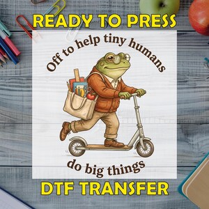 May include: A DTF transfer featuring a green frog wearing glasses, a brown cardigan, and tan trousers, riding a scooter while carrying a tote bag filled with school supplies. The text reads READY TO PRESS, Off to help tiny humans do big things.