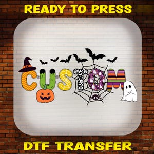 May include: A Halloween-themed DTF transfer graphic featuring the word CUSTOM in stylised letters. The letters incorporate orange chevron, green monster, yellow spiderweb, black pumpkin, purple ghost, and striped patterns with bats and a ghost.