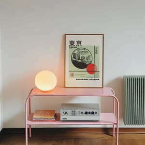 Op de afbeelding: Een roze metalen bijzettafel met twee planken. De bovenste plank heeft een witte globe lamp en een vintage stereo-installatie. De onderste plank heeft een stapel boeken. Een ingelijst poster met de tekst "Tokyo" en "Shiodome Station" hangt aan de muur achter de tafel.