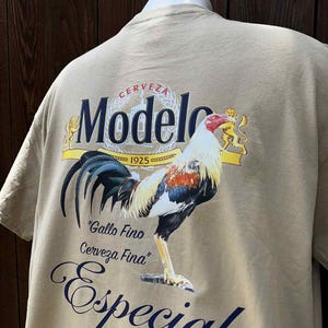 May include: Tan t-shirt featuring a colorful rooster graphic with the words "Modelo" and "Especial." The rooster has red, white, and orange feathers. The shirt also includes the text "Gallo Fino Cerveza Fina."