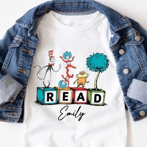 May include: White t-shirt featuring a colorful illustration of characters from a children's book. The word "READ" is spelled out on blocks, with the name "Emily" below. A denim jacket is draped over the shirt.