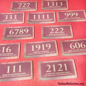 May include: A collection of rectangular cards with various number combinations and corresponding messages in white text. The cards have a purple background with a subtle glitter effect and a white border. The cards are arranged on a pink surface.