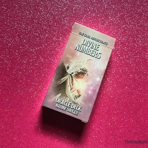 May include: A rectangular oracle deck titled "Divine Numbers" by Nadine Swiger, featuring an angel illustration. The deck is set against a sparkly pink background. The text "The Soul Reflection's" is at the top.