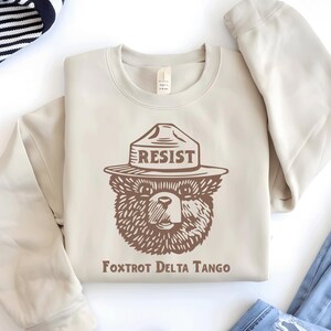 Pu&ograve; includere: Una felpa beige con una grafica marrone di un orso che indossa un cappello da ranger con la scritta "RESIST" su di esso. Il testo "Fox Trot Delta Tango" &egrave; stampato sotto l'orso.