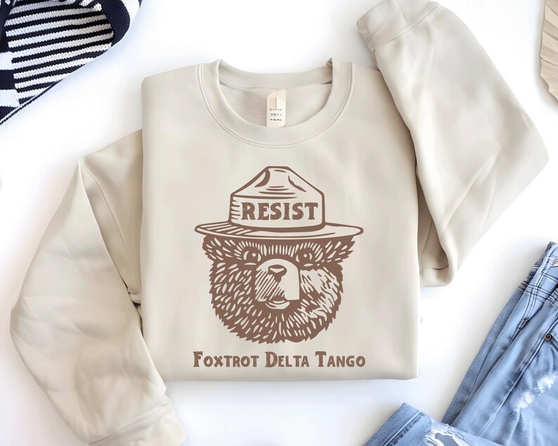 Pu&ograve; includere: Una felpa beige con una grafica marrone di un orso che indossa un cappello da ranger con la scritta "RESIST" su di esso. Il testo "Fox Trot Delta Tango" &egrave; stampato sotto l'orso.