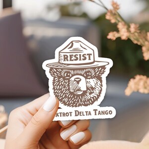 Pu&ograve; includere: Un orso bruno che indossa un cappello da ranger con la scritta "RESIST" sopra. L'adesivo ha anche il testo "XtroT Delta Tango" in basso.