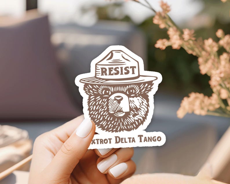 Pu&ograve; includere: Un orso bruno che indossa un cappello da ranger con la scritta "RESIST" sopra. L'adesivo ha anche il testo "XtroT Delta Tango" in basso.