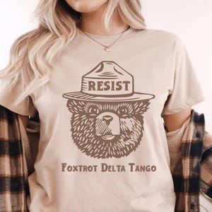 Pu&ograve; includere: Una maglietta beige con un'immagine di un orso che indossa un cappello da ranger con la scritta "RESIST" su di esso. Il testo "FOXTROT DELTA TANGO" &egrave; stampato sotto l'immagine.