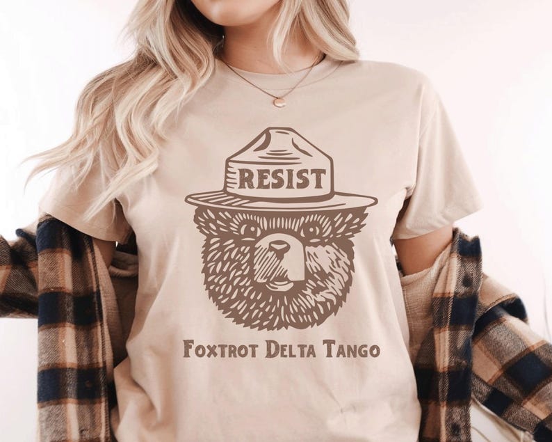 Pu&ograve; includere: Una maglietta beige con un'immagine di un orso che indossa un cappello da ranger con la scritta "RESIST" su di esso. Il testo "FOXTROT DELTA TANGO" &egrave; stampato sotto l'immagine.
