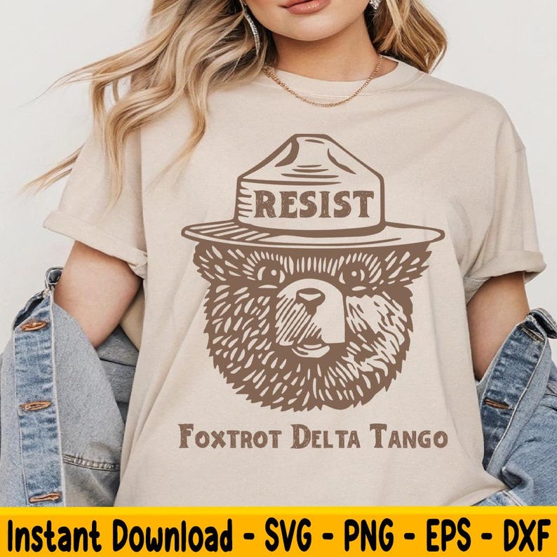 Pu&ograve; includere: Una maglietta beige con un orso marrone che indossa un cappello da ranger con la scritta "Resist". Il testo "Foxtrot Delta Tango" &egrave; sotto l'orso.