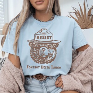 Pu&ograve; includere: Maglietta blu chiaro con un orso di cartone marrone che indossa un cappello da ranger che dice "Resist". Il testo "Fox Trot Delta Tango" &egrave; stampato sotto l'orso.