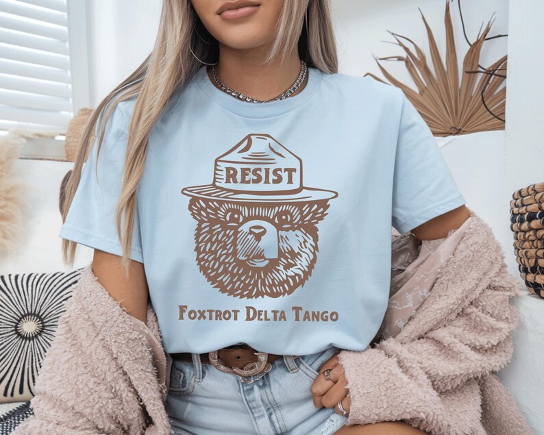 Pu&ograve; includere: Maglietta blu chiaro con un orso di cartone marrone che indossa un cappello da ranger che dice "Resist". Il testo "Fox Trot Delta Tango" &egrave; stampato sotto l'orso.