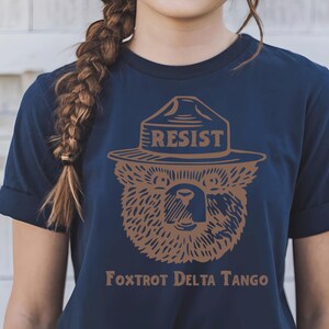 Pu&ograve; includere: Una maglietta blu navy con un'immagine di un orso marrone che indossa un cappello da ranger con la scritta "RESIST" sopra. Il testo "FOXTROT DELTA TANGO" &egrave; stampato sotto l'orso.