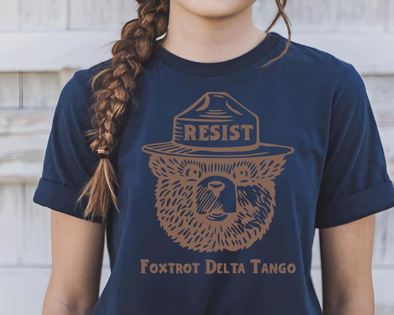 Pu&ograve; includere: Una maglietta blu navy con un'immagine di un orso marrone che indossa un cappello da ranger con la scritta "RESIST" sopra. Il testo "FOXTROT DELTA TANGO" &egrave; stampato sotto l'orso.