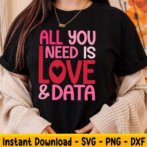 Peut inclure: Un t-shirt noir avec le texte "All you need is love & data" en lettres roses et rouges. Le texte est dans une police de caractères ludique et comprend un cœur rose.