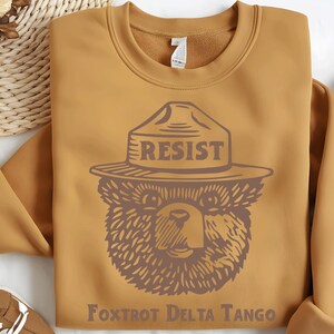 Pu&ograve; includere: Una felpa color giallo senape con una grafica di un orso che indossa un cappello da ranger con la scritta "RESIST" su di esso. Il testo "FOXTROT DELTA TANGO" &egrave; stampato sotto l'orso.