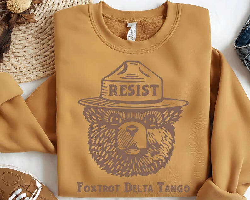 Pu&ograve; includere: Una felpa color giallo senape con una grafica di un orso che indossa un cappello da ranger con la scritta "RESIST" su di esso. Il testo "FOXTROT DELTA TANGO" &egrave; stampato sotto l'orso.