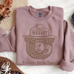 Pu&ograve; includere: Una felpa color malva con una grafica marrone di un orso che indossa un cappello da ranger con la scritta "RESIST" su di esso. Il testo "Foxtrot Delta Tango" &egrave; stampato sotto l'orso.