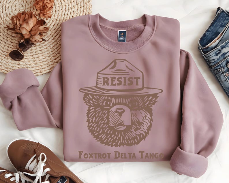 Pu&ograve; includere: Una felpa color malva con una grafica marrone di un orso che indossa un cappello da ranger con la scritta "RESIST" su di esso. Il testo "Foxtrot Delta Tango" &egrave; stampato sotto l'orso.