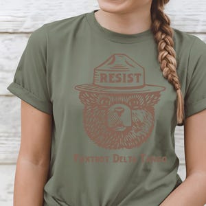Pu&ograve; includere: Una maglietta verde con un orso marrone che indossa un cappello da ranger con la scritta "Resist". Il testo "Fox Trot Delta Tango" &egrave; stampato sotto l'orso.