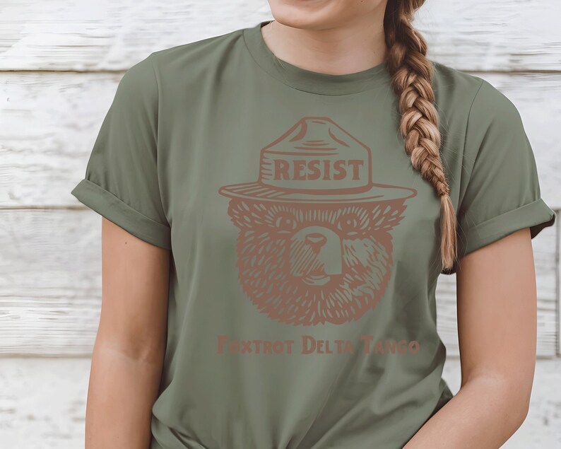 Pu&ograve; includere: Una maglietta verde con un orso marrone che indossa un cappello da ranger con la scritta "Resist". Il testo "Fox Trot Delta Tango" &egrave; stampato sotto l'orso.