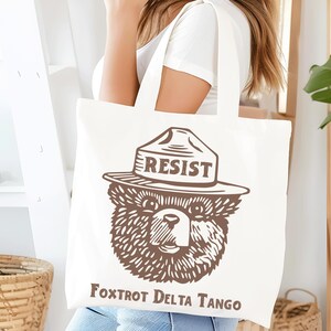 Pu&ograve; includere: Borsa tote in tela bianca con un'illustrazione marrone di un orso che indossa un cappello da ranger con la scritta "RESIST" su di esso. Il testo "Fox Trot Delta Tango" &egrave; stampato sotto l'orso.