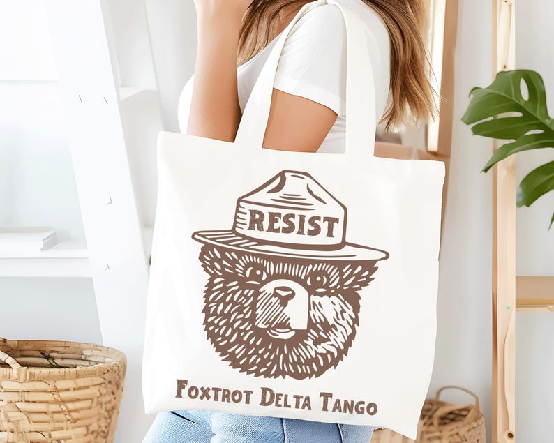 Pu&ograve; includere: Borsa tote in tela bianca con un'illustrazione marrone di un orso che indossa un cappello da ranger con la scritta "RESIST" su di esso. Il testo "Fox Trot Delta Tango" &egrave; stampato sotto l'orso.