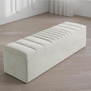 May include: A rectangular, light-colored storage bench with a padded, ribbed top. The bench is upholstered in a textured fabric and sits on a wooden floor. The design is simple and modern, suitable for various interior styles.