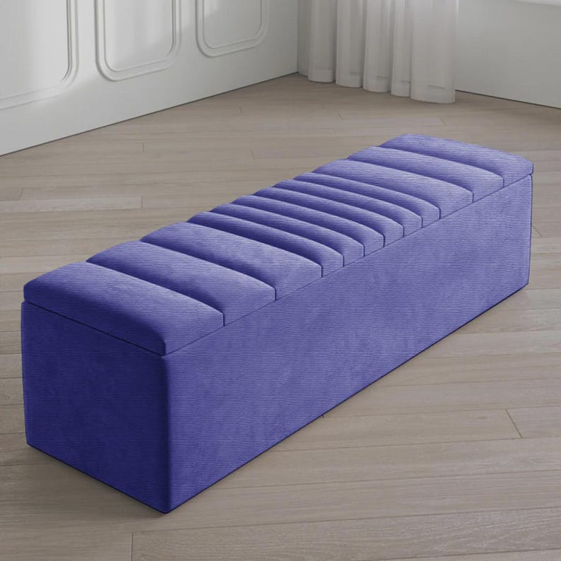 May include: A long, rectangular storage bench in a vibrant periwinkle blue. The bench features a padded, ribbed top and a solid, boxy base. The fabric has a subtle texture, and the bench is set on a light-colored wooden floor.