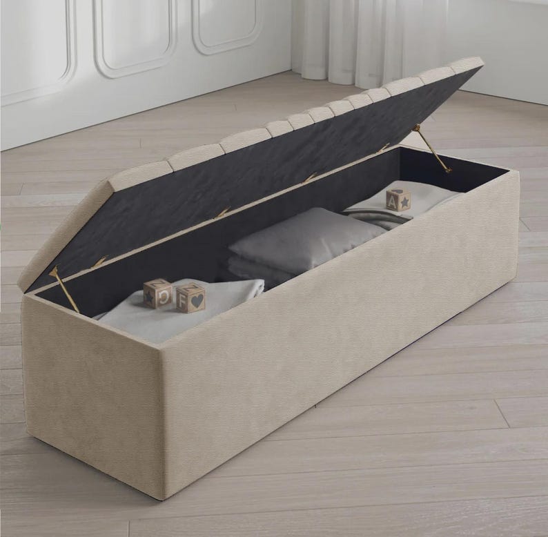 May include: A beige upholstered storage bench with a hinged lid, revealing a dark interior. Inside are pillows, blankets, and wooden blocks. The bench is rectangular and sits on a light-colored wooden floor.