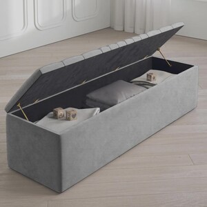 May include: A gray upholstered storage bench with the lid partially open, revealing a dark interior. Inside are folded blankets, a pillow, and wooden blocks. The bench is rectangular and sits on a light-colored wooden floor.
