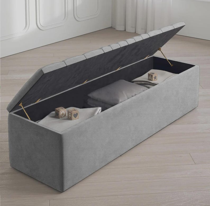 May include: A gray upholstered storage bench with the lid partially open, revealing a dark interior. Inside are folded blankets, a pillow, and wooden blocks. The bench is rectangular and sits on a light-colored wooden floor.