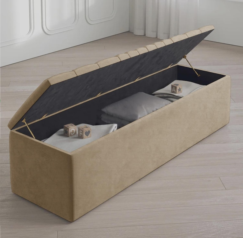 May include: A beige rectangular storage bench with a hinged lid, partially open to reveal folded blankets and pillows. The interior is black, and the lid's top has a tufted design. Wooden blocks with letters and symbols are inside.