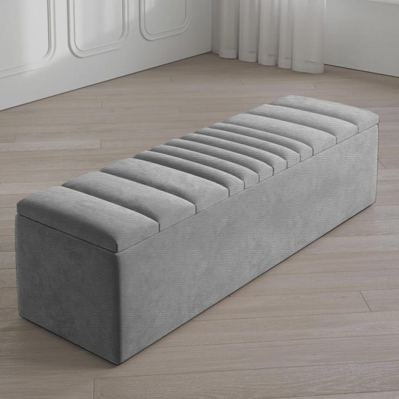 May include: A gray upholstered storage bench with a rectangular shape. The bench features a padded, ribbed top and a solid base. The neutral color and simple design make it suitable for various interior styles. The bench is approximately 60 inches long.