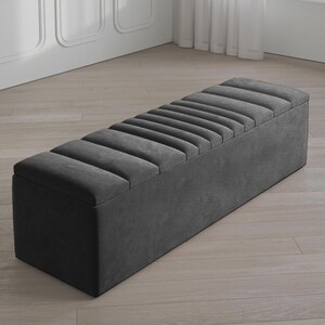 May include: A dark gray upholstered storage bench with a rectangular shape. The top features a series of parallel, padded sections. The bench is placed on a light-colored wooden floor, and the background includes a white wall and a window.